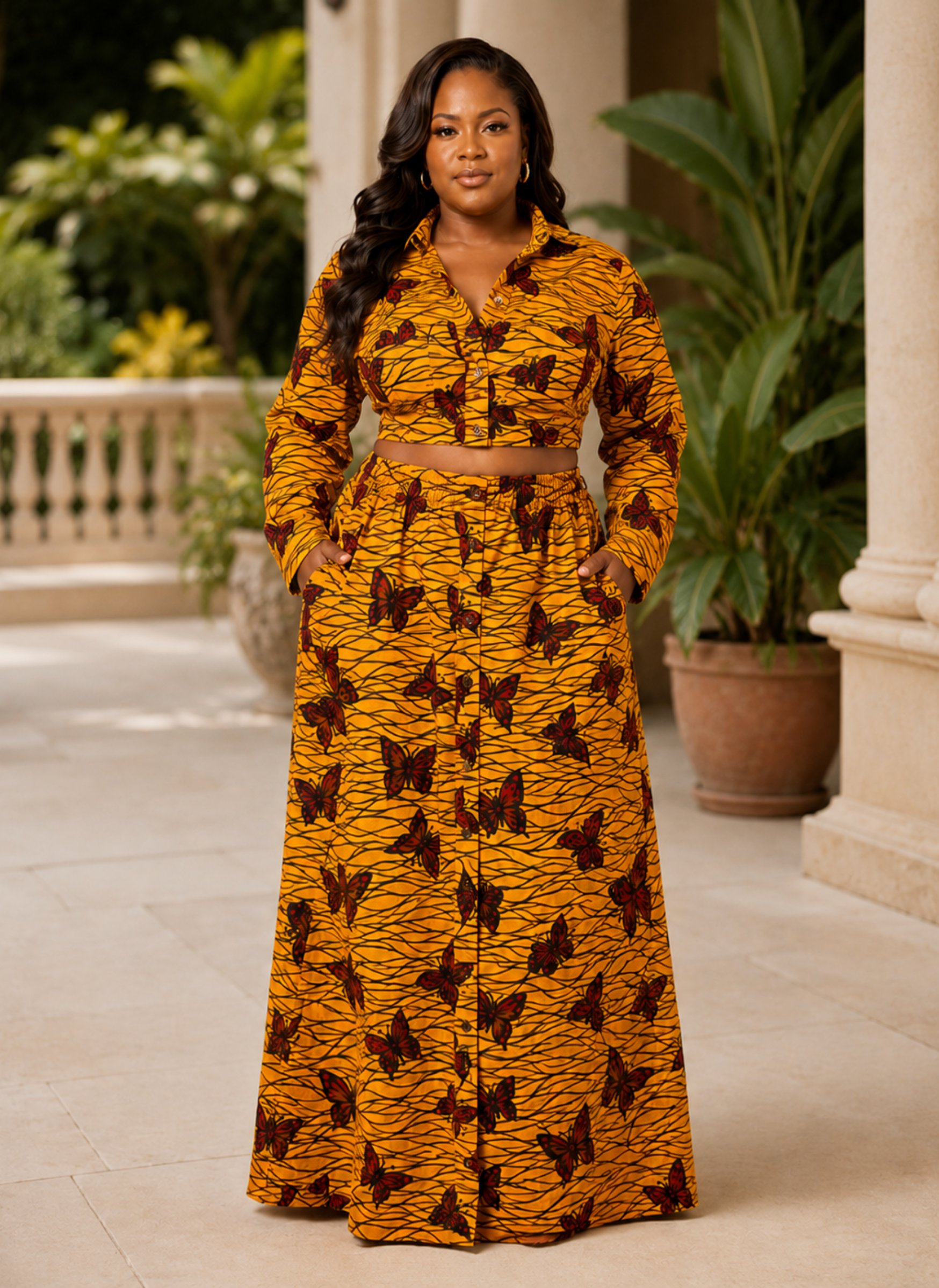 A woman stands on a sunlit outdoor porch wearing a matching long-sleeve button-front top and high-waisted maxi skirt set in a mustard yellow fabric with a dark brown butterfly print and a thin brown waist tie.