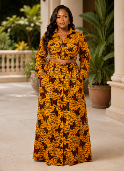 A woman stands on a sunlit outdoor porch wearing a matching long-sleeve button-front top and high-waisted maxi skirt set in a mustard yellow fabric with a dark brown butterfly print and a thin brown waist tie.