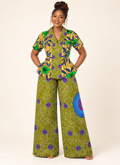 A woman modeling a matching two-piece outfit featuring a short-sleeve peplum top and wide-leg pants in a vibrant green, yellow, blue, and purple African wax print pattern, set against a plain beige studio background.