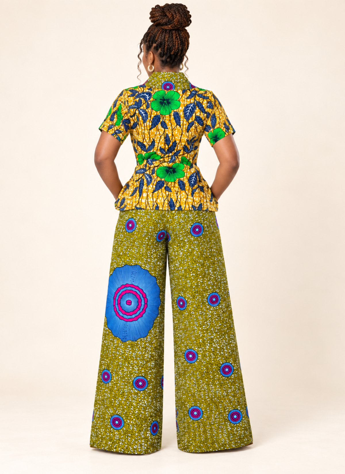 A woman shown from the back wearing a matching two-piece outfit with a short-sleeve peplum top and wide-leg pants in a vibrant yellow, green, blue, and purple African wax print pattern, set against a plain beige studio background.