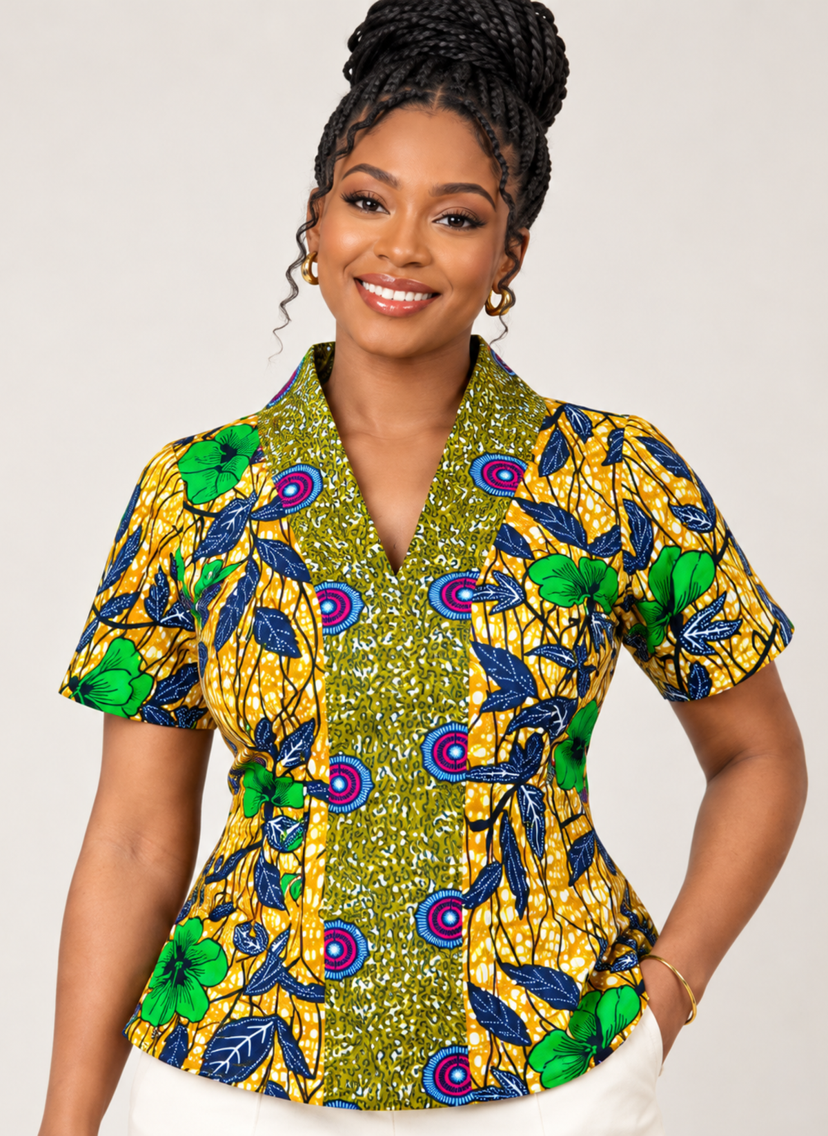 A woman wearing a short-sleeve collared button-up shirt with a vibrant African wax print pattern in yellow, green, blue, and purple, paired with white pants against a light gray studio background.