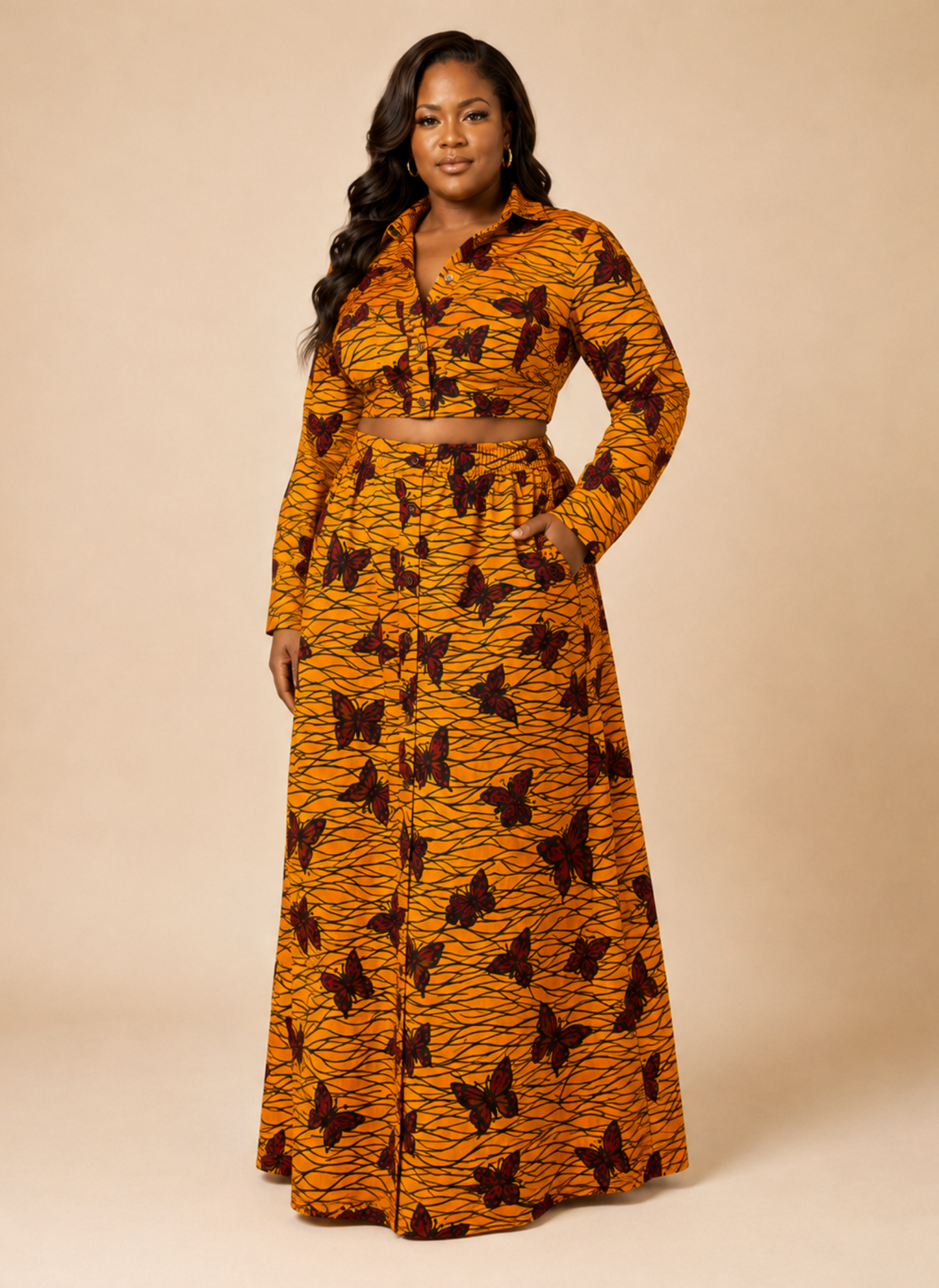 A woman modeling a matching long-sleeve crop top and maxi skirt set in a vibrant orange and black butterfly print, set against a solid beige studio background.