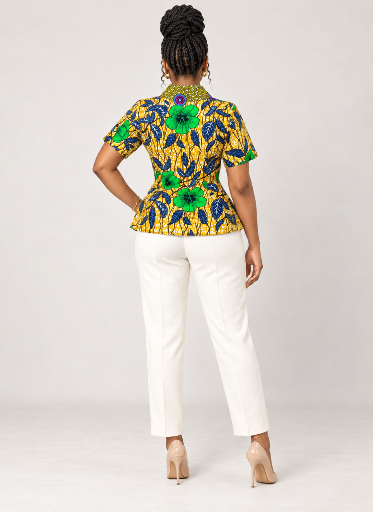 A woman shown from the back wearing a short-sleeve peplum-style top with a bold yellow, green, and blue floral print, paired with white pants and beige heels, against a light gray studio background.