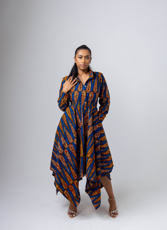 A model wearing a long sleeve shirt dress with an asymmetric hem, featuring a geometric pattern in shades of blue and orange. The dress has buttons and pockets, and is possibly tied at the waist with a belt.
